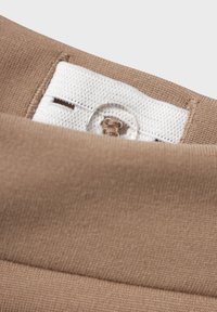 Brauner Stoff mit glatter Textur, der ein weißes Etikett mit Knopfloch und Nähdetail aufweist. Sichtbare Naht und Farbvariationen.