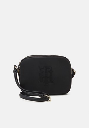 Schwarze rechteckige Umhängetasche mit eingeprägtem Logo auf der Vorderseite, goldfarbenen Beschlägen und verstellbarem Riemen, der neben der Tasche aufgerollt liegt.