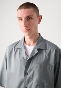 Camisa gris de manga corta con cuello extendido, bolsillo frontal y cierre de botones. Textura suave con un corte relajado. Acompañada de una camiseta blanca por debajo.