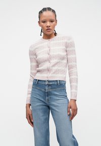 Jauna sieviete ar pītām matiem, tērpusies rozā un baltās svītrainās pogu jakās un augsta jostasvietā zilos džinsos uz baltas fona.