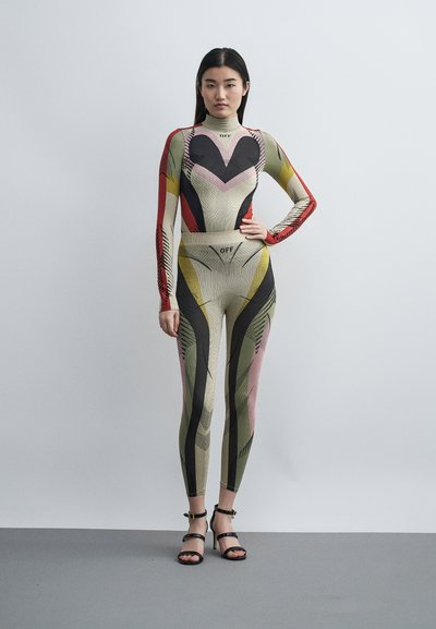 Γυναίκα στέκεται με το χέρι στη μέση, φοράει πολύχρωμη, μοτίβο εφαρμοστό ολόσωμο φόρεμα και μαύρα σανδάλια με ψηλό τακούνι σε απλό γκρι φόντο.