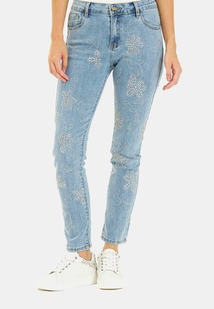 Light blue denim jeans with silver rhinestone floral patterns, five pockets, and a slim fit, paired with white sneakers featuring embellishments.