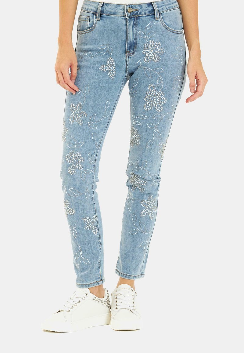 Jeans in denim azzurro chiaro con motivi floreali in strass argento, cinque tasche e una vestibilità slim, abbinati a sneakers bianche con decorazioni.