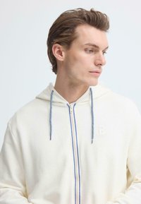 Sweat à capuche crème zippé avec un tissu texturé, des cordons bleus et un logo discret sur le côté gauche.