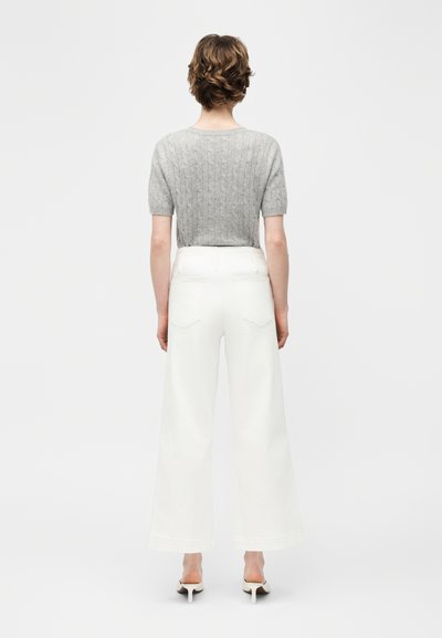 Άτομο με κοντά καστανά μαλλιά, φορώντας γκρι κοντομάνικη πλεκτή μπλούζα, λευκό παντελόνι φαρδιάς γραμμής και λευκά παπούτσια με ψηλό τακούνι, στέκεται γυρισμένο με την πλάτη.