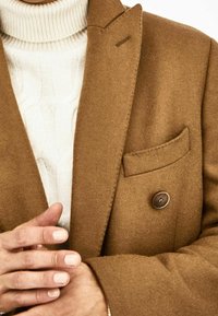 Brauner Wollmischung Doppelreiher mit Reverskragen, ausgestattet mit einer Brusttasche und einem einzelnen Knopf. Darunter ist ein cremefarbener, kabelgestrickter Rollkragenpullover sichtbar.