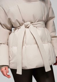 Beige prošívaná bunda s širokým páskem uvázaným na uzel v pase, nošena přes černé legíny, ruce uvolněně po stranách.