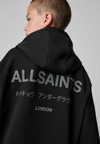 Deček, ki gleda vstran, oblečen v črn kapuco s tiskom "ALLSAINTS," japonskim besedilom in "LONDON" na hrbtni strani, na enobarvni sivi podlagi.