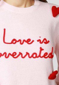 Maglione rosa chiaro lavorato a maglia con scritta ricamata rossa "Love is overrated" e cuori rossi decorativi sulle spalle.