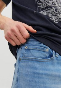 Left hand pulling up navy blue graphic t-shirt over light blue denim jeans pocket on light background.