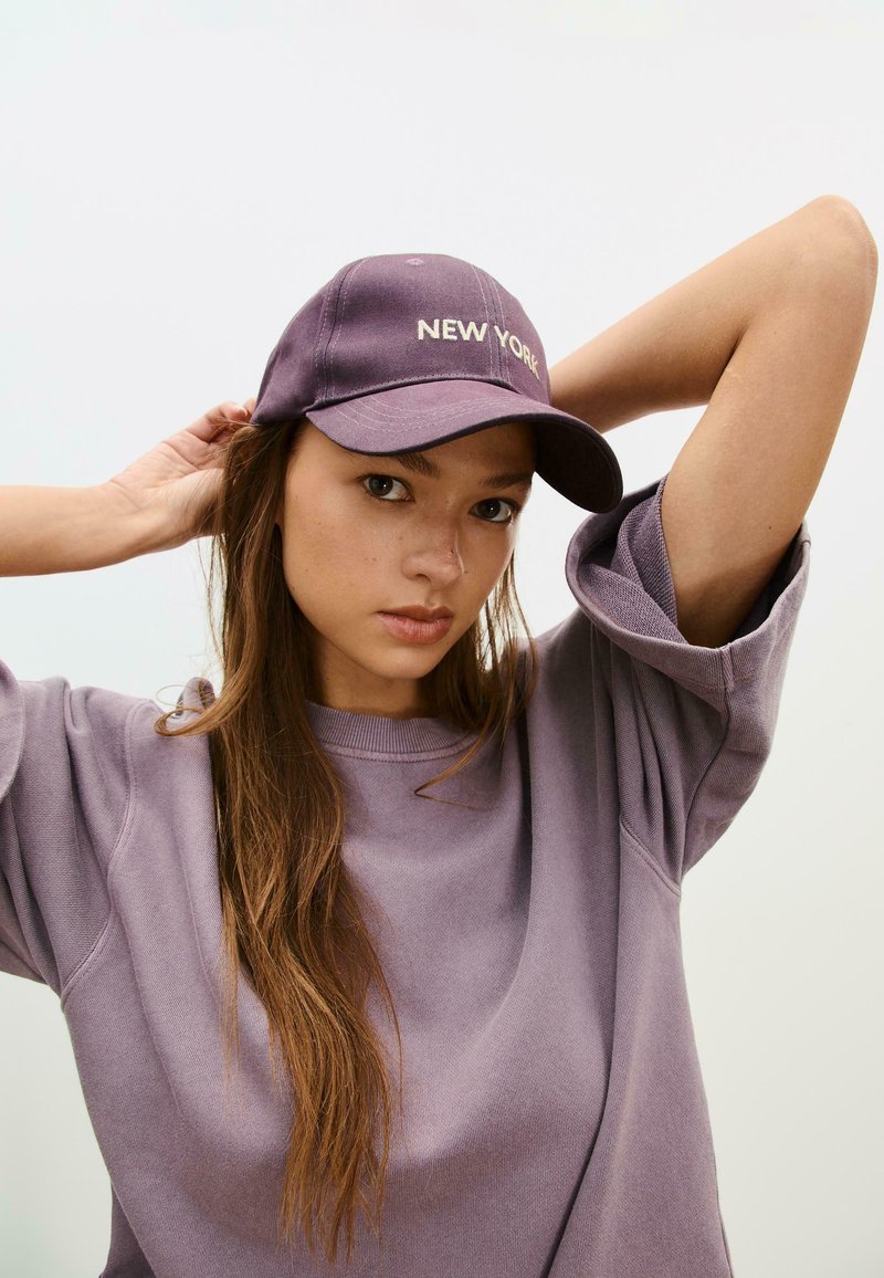 Lila basebollkeps med "NEW YORK" broderat i ljusgult, bärs med en matchande överstor lila sweatshirt i texturerat tyg.