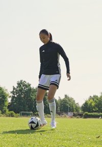 Schwarze Sportjacke mit weißen Akzenten, weiße Shorts mit schwarzen Streifen, steht auf Gras und bereitet sich darauf vor, einen schwarz-weißen Fußball zu kicken.