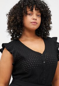 Black dotted blouse with V-neck, button closure, and ruffled short sleeves. Textured fabric features small cutouts in a patterned design.