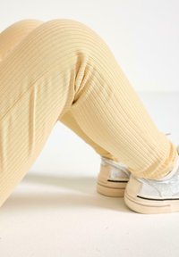 Lys gule ribbede leggings med en blød tekstur, parret med sølvglitrende sneakers med en kontrasterende sort stribe på en cremefarvet overflade.
