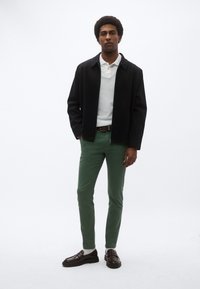 Μαύρο υφασμάτινο σακάκι, λευκό πόλο πουκάμισο, πράσινο παντελόνι στενής γραμμής και καφέ μοκασίνια με παχιές σόλες. Στέκεται μπροστά σε λευκό φόντο.