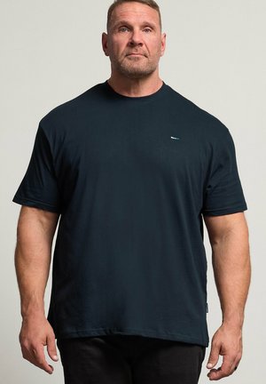 Muskeløs mand med kort hår og trimmet skæg står vendt fremad, iført en ensfarvet sort T-shirt med korte ærmer og sorte bukser.