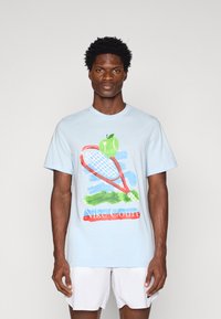 Svetlo modré bavlnené tričko s grafikou tenisového rakety a zelenej jablka, s textom "Nike Court" v červenej farbe. Krátke rukávy, voľný strih.