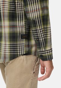 Close-up van een persoon in een groen en zwart geruit shirt met een label "Made for Daily Adventures", gecombineerd met beige corduroybroek.