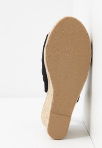 Semelle d'une sandale espadrille beige à talon compensé avec semelle en caoutchouc texturée et dessus en tissu noir, positionnée contre un mur et un sol blancs.