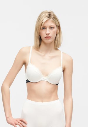 Mujer rubia con sujetador acolchado blanco con ribete de encaje y braguitas blancas de talle alto, de pie contra un fondo claro y liso.