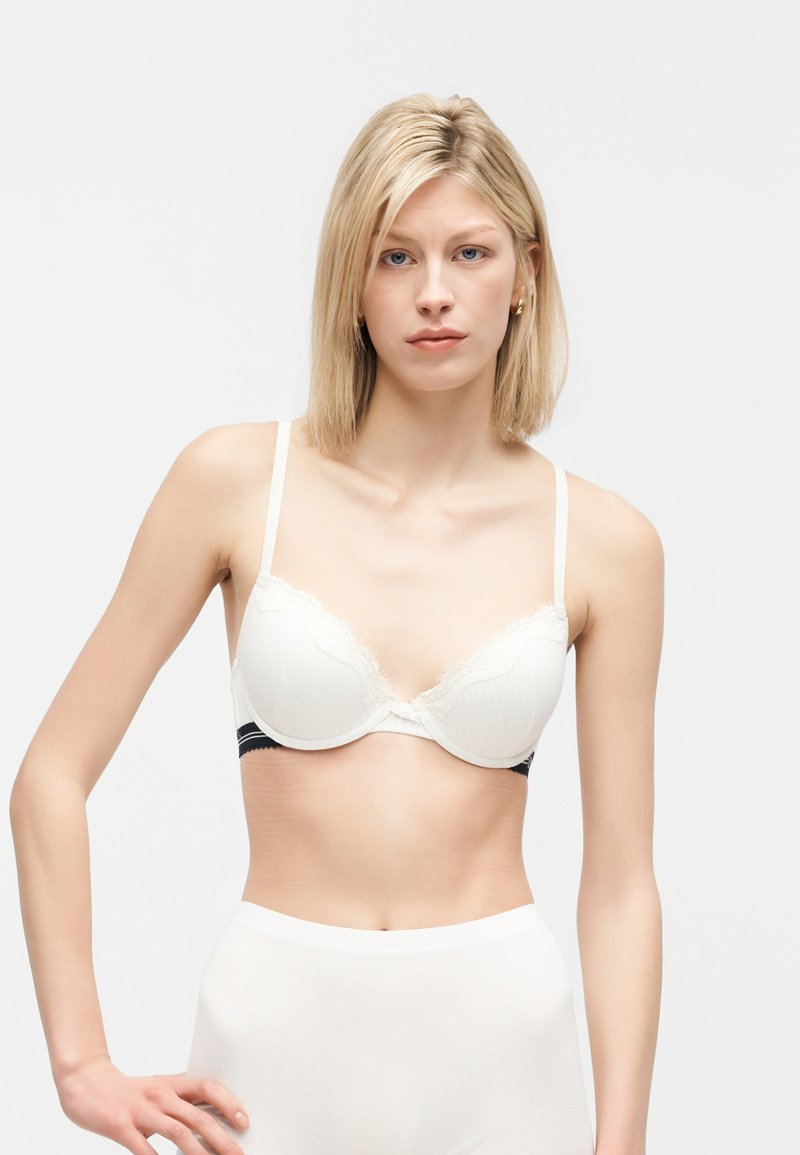 Lyshåret kvinde iført hvid polstret bh med blonder og hvide højttaljede trusser, stående mod en ensfarvet lys baggrund.