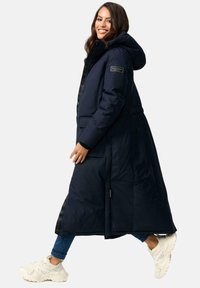 Donker marineblauwe pufferjas met capuchon, elastische tailleband en zijzakken. Bevat een subtiel logo op de mouw en heeft een gladde buitenkant.