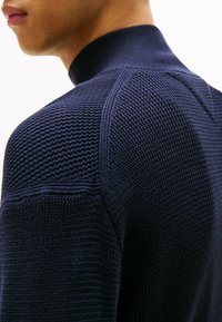 Pull en tricot bleu marine avec un col montant, présentant des motifs texturés et des accents côtelés sur les épaules et les manches.