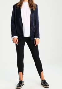 Blazer blu navy con una trama liscia, camicetta bianca plissettata, leggings neri con fessure laterali e mocassini neri lucidi.