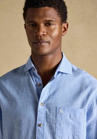 Mann trägt ein hellblaues Hemd mit Knöpfen, einer Brusttasche und einem kleinen eingestickten Logo vor einem schlichten beigen Hintergrund.