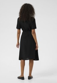 Robe noire, longueur genou, avec des manches courtes, une taille cintrée et une jupe fluide. Tissu lisse, peu de détails, design simple.