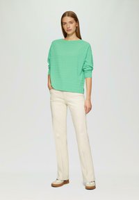 Camicia verde a maniche lunghe con texture abbinata a pantaloni beige a gamba dritta e sneakers color crema. L'outfit presenta una vestibilità rilassata e un design minimalista.