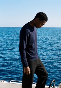 Jeune homme debout sur un bateau, regardant vers le bas, avec une mer bleue calme et un ciel clair en arrière-plan.