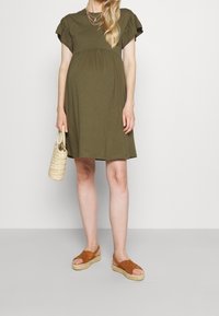 Zielona sukienka ciążowa z krótkim rękawem, dopasowaną górą i rozkloszowanym dołem. Noszona z brązowymi espadrylami i plecioną torebką.