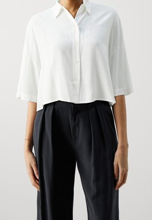 Femme portant une chemise blanche ample boutonnée avec des manches mi-longues et un pantalon plissé noir taille haute.