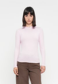Lichtroze geribbelde longsleeve top met een golvende halslijn en een strakke pasvorm, gecombineerd met bruine broek.