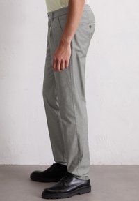 Mann in grauen Maßanzug-Hosen und schwarzen Dressschuhen, der gegen eine hell strukturierte Wand steht.