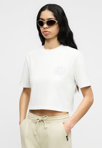 Γυναίκα με μακριά σκούρα μαλλιά που φοράει μπεζ γυαλιά ηλίου με σχήμα cat-eye, λευκό κοντομάνικο μπλουζάκι με λεπτό σχέδιο στο στήθος και μπεζ παντελόνι με κορδόνι, τα χέρια της στις τσέπες.