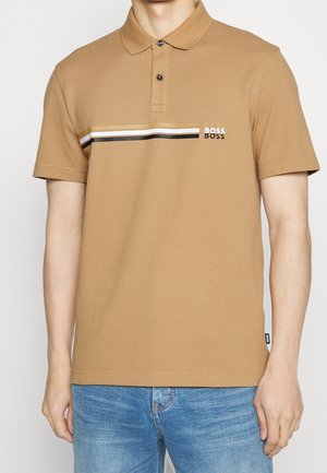 Mand iført en beige poloshirt med sorte, hvide og beige vandrette striber og ordet BOSS på brystet, kombineret med blå jeans.