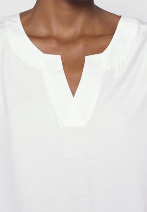 Personne à la peau foncée portant un haut blanc avec un large décolleté en V et une petite encoche au centre.