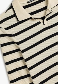 Camisa polo rayada en beige y negro con textura acanalada y un cuello clásico. El diseño presenta rayas alternas de diferentes anchos.