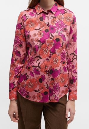 Femme portant une chemise rose à boutons avec un motif de grandes fleurs orange et violettes, associée à un pantalon marron.