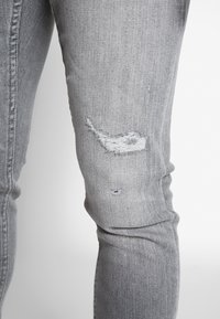 Šedé deninové džíny s slim fit střihem, s opotřebenými místy na kolenou, odkryvajícími strukturu spodní látky a malými dírami.