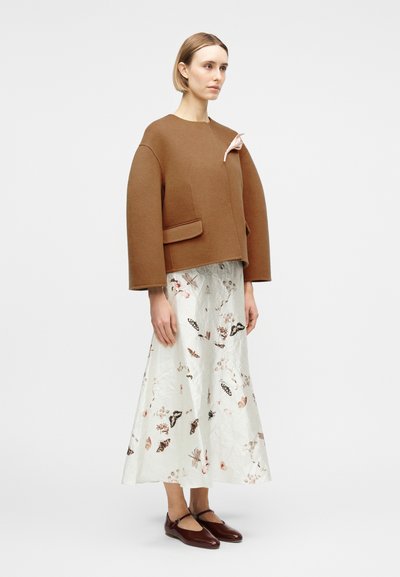 Femme portant une veste marron oversize à larges manches, une longue jupe blanche à motifs floraux, et des chaussures Mary Jane marron, debout devant un fond uni.