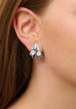 Silver metal earrings featuring a geometric, abstract triangular design with a polished finish, worn on the lobe of an ear.