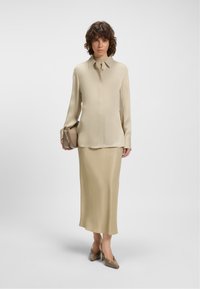 Woman standing wearing beige long-sleeve blouse, matching long skirt, carrying a beige handbag, and wearing beige heels on a white background.