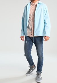 Hellblaue wasserdichte Jacke mit verstellbarem Saum, getragen über einem gemusterten rosa Hemd. Paar zerrissene blaue Jeans und graue Turnschuhe.