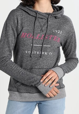 Personne portant un sweat à capuche Hollister gris foncé avec des lettres roses et blanches et un jean bleu clair, les mains posées sur la taille.