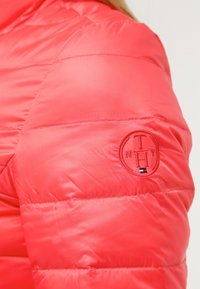 Vadderad röd jacka med en texturerad yta, horisontell sömnad och en cirkulär logotyp på ärmen med initialer och en liten flagga som accent.