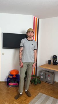 Siva majica s kratkim rukavima s crnim obrubom, maslinaste hlače, smeđe cipele i tetovaže. Unutarnji prostor s šarenom zidnom akcentom i dekorom.