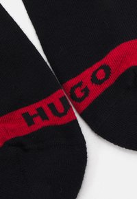 Schwarze Socken mit glatter Textur zeigen ein auffälliges rotes Band, auf dem am oberen Rand "HUGO" in schwarzer Schrift zu sehen ist.